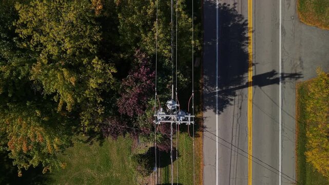 Aerial drone view of an electric power line pole casting a long shadow on a rural asphalt road in the United States surrounded by autumn trees.