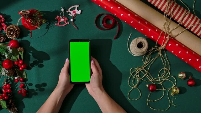 Top view of female hands holding a smartphone with a green screen on a dark green festive background. Holiday wrapping materials, ribbons, gold beads, red ornaments, and pine decorations surround the