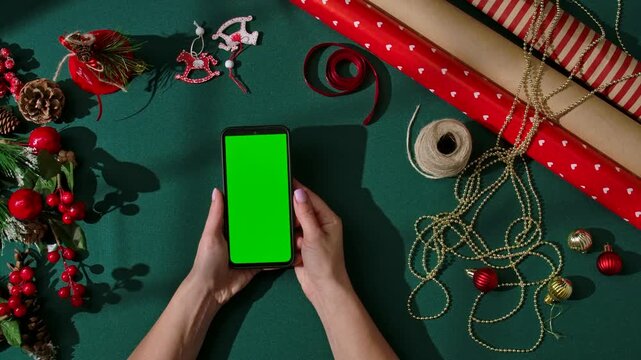 Top view of female hands holding a smartphone with a green screen on a dark green festive background. Holiday wrapping materials, ribbons, gold beads, red ornaments, and pine decorations surround the