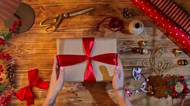 Top view of a woman hands tying a red satin ribbon on a gift box among scissors, twine, and decorations on a rustic wooden table for Christmas crafts.