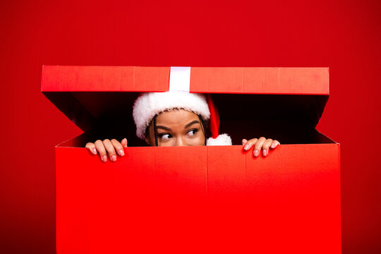 A playful young woman in a Santa hat peeks out of a giant red gift box against a bright red background creating a funny festive moment