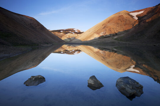 View of the serene lake mirrors the rugged, sunlit mountains, with rocks emerging from the calm water, creating a striking contrast of textures and colors, Landmannalaugar, Rang&aacute;rping ytra, Iceland.