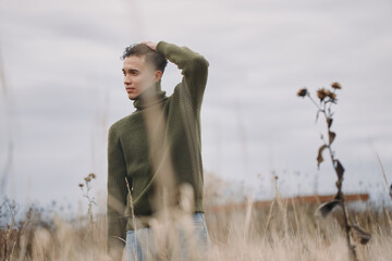 Young man in a green knit sweater standing in a dry field, contemplative gaze conveying...