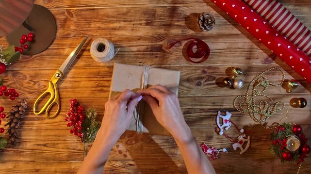 Top view of a woman hands adjusting a gift wrapped in kraft paper, tied with twine, decorated with fir branches and red berries, on a rustic wooden table using scissors and ribbon. Cozy handmade