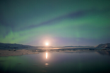View of the ethereal aurora borealis dances above the icy lagoon, its green tendrils mirroring the moonlight on the still water, Jokulsarlon, Sveitarfélagio Hornafjorour, Iceland.
