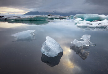View of translucent icebergs drift serenely on the tranquil, reflective waters under a cloudy sky, with distant mountains, Jokulsarlon, Sveitarfélagio Hornafjorour, Iceland.