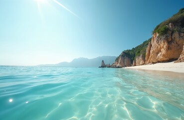 Fototapeta premium Clear turquoise ocean water laps onto a sandy beach with rugged cliffs and green vegetation under a bright sunny sky. Peaceful coastal landscape invites relaxation and adventure.