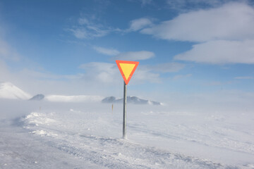 View of a yellow and red triangular road sign standing starkly against a backdrop of snow-covered terrain and a pale blue sky, Iceland, Iceland.