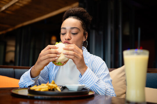 A happy young African American woman enjoying a delicious burger with fries at a modern restaurant - Powered by Adobe