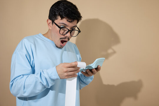 Man with glasses holding a bill and a blue calculator, looking surprised with his mouth open on a light brown background, concept of unexpected expenses and personal finance