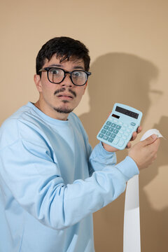 Man with glasses holding a blue calculator and a bill, looking worried on a light brown background with copy space, concept of debt and personal finance
