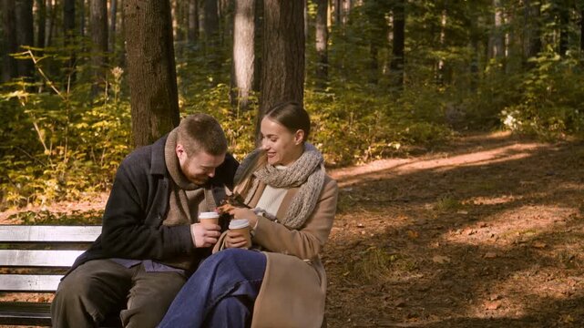 Long copy space shot of young Caucasian couple kissing while drinking coffee sitting on bench with adorable small dog in fall park