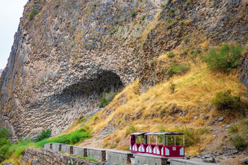 A stunning view of the Symphony of Stones basalt cliff in Garni Gorge, Armenia, with a small red and white tourist train passing below. The dramatic hexagonal rock formations, shaped by volcanic