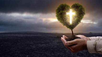 Human hand holding light of the Christian cross on the heart tree with barren field and dark dramatic sky background. Concept of Christianity, religious, faith, Jesus, or belief
