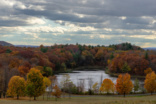 View of autumn colors painting the dense forest surrounding a tranquil lake under a dramatic, cloudy sky, Poughkeepsie, New York, United States.