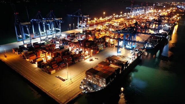 Aerial view of a cargo ship moored at a busy dock illuminated at night for unloading containers using large cranes.