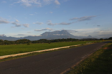 Road to Mount Kinabalu View. A paved, empty road leads the eye toward the distant, majestic Mount Kinabalu framed by green fields and a blue, cloudy sky in Sabah, Malaysia. Represents travel and desti