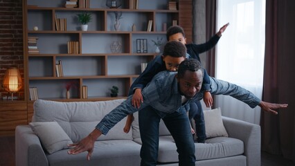 Carefree African American family playing piggyback pretending airplane at home joyful father dad riding on back little son play plane flying pretend with two kids boys sons children having fun joy