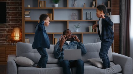 African American family irritated father work noisy kids jump on couch dad with laptop talking mobile phone annoyed busy near two children sons little boys jumping on sofa parenthood trouble stress