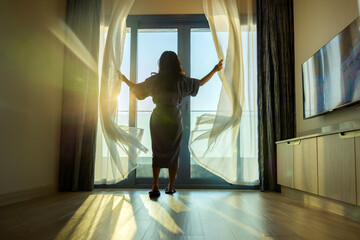 Excited happy woman opens the curtains white waving curtain, relaxes in sunlight, enjoying a...