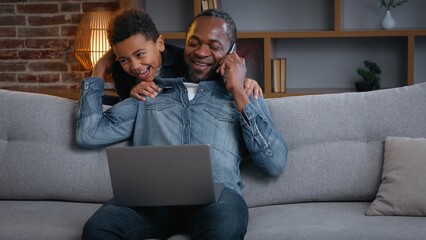Happy African American family father multitasking son cuddling dad surprise dad working with laptop talking mobile phone call at home distant job little boy kid child hugging daddy fun love hug cuddle