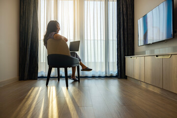 Middle aged woman with Blank screen laptop for presentation media of business watching TV. Sunlight through sheer curtains, casting shadows on wooden floor. The modern room creating serene atmosphere.
