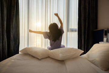 Good morning, New day, Holiday. Happy asian middle aged woman wake up on bed, stretching her arms in fresh early morning, relax peaceful morning routine. Luxury hotel or home in the morning sunlight.