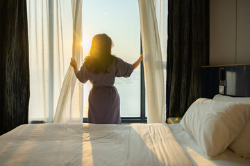 Healthy sleep, Recreation and Relaxation Concept. Happy middle aged woman waking up in bedroom and opens the curtains to let the Soft morning sunlight streaming through window. Greeting new day.