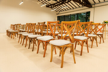 Rows of wooden chairs with white cushions arranged for conference, seminar, or wedding ceremony in modern event hall with natural lighting and wooden ceiling beams
