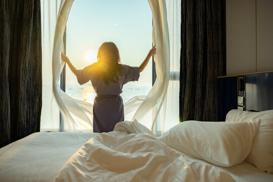 Confident middle aged woman opens the curtains with white flying curtains, looking at the view from window Beautiful sunrise above sea or ocean. Woman enjoying her holiday in a luxury hotel. Back view