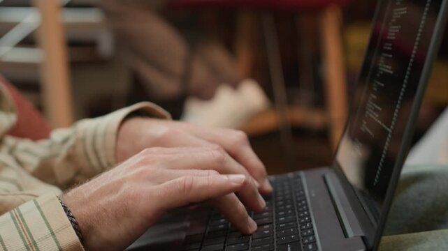 Rack focus shot of hands of unrecognizable programmer coding on laptop sitting in modern coworking space with his male colleague sitting at desk and typing on laptop