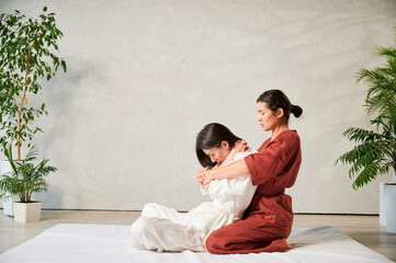 Masseuse making thai yoga massage. Woman therapist doing traditional massage treatment, stretching female patient's body. Client sitting on mat, enjoying hand massage in calm atmosphere.
