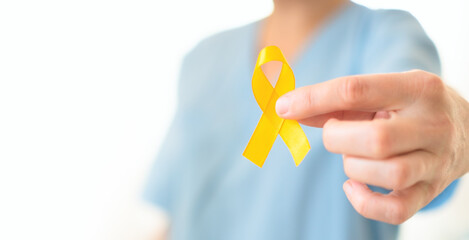 Close up of female doctor and nurse in uniform holding yellow ribbon in hand. Horizontal banner. Symbol of childhood cancer fight, medical support, diagnosis, treatment, family care, and awareness.