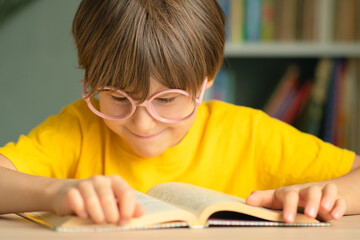 Curious happy child with glasses sits at desk, leaning on hand and looking at open book. Concept of reading, childhood study, learning, early education, preparation for school and knowledge.