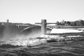 A black and white view of the Rainbow Bridge and Niagara Falls Observation Tower rising above mist...