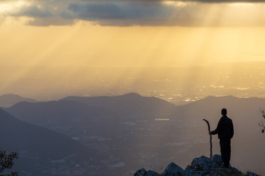 View of a lone figure standing on a rocky precipice, staff in hand, gazing over the misty, sun-drenched valleys and distant city lights, Pizzo San Michele, Campania, Italy.