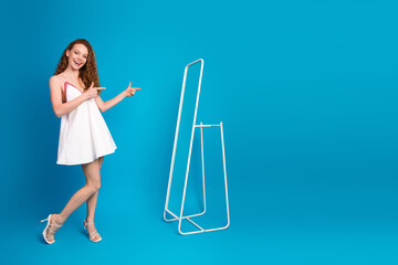 Young woman in a white dress stands by a minimal metal rack against a bright blue background smiling and pointing playfully at the scene creating a fun fashionable lifestyle moment