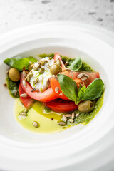 A gourmet burrata salad with fresh sliced tomatoes, basil, green olives, and sunflower seeds, drizzled with basil pesto and olive oil in a white bowl on a light background