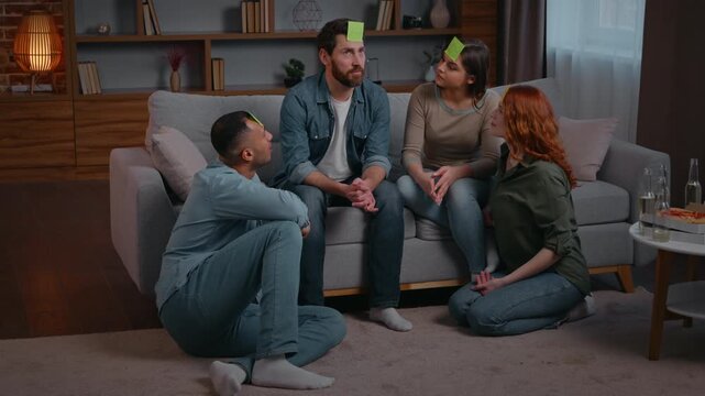Four diversity ethnic friends playing guessing game stickers on head at home party funny leisure multiracial women men in living room playing charades talking communicate asking Who am I play joyful