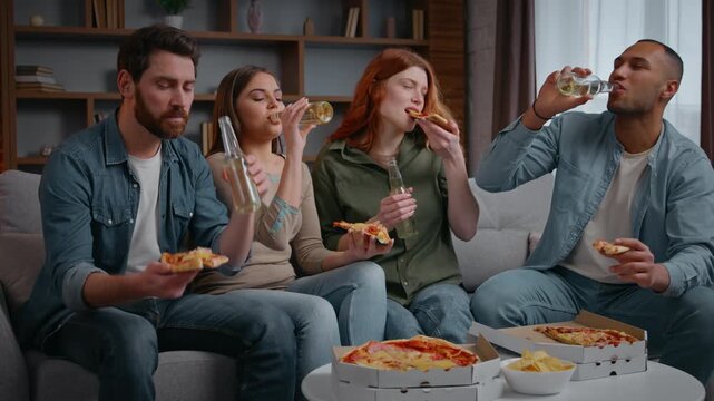 Happy multiracial four friends home party clinking bottles beer alcohol eating pizza on couch diversity buddies cheers with drinks talking communicate friendship birthday event celebration men women
