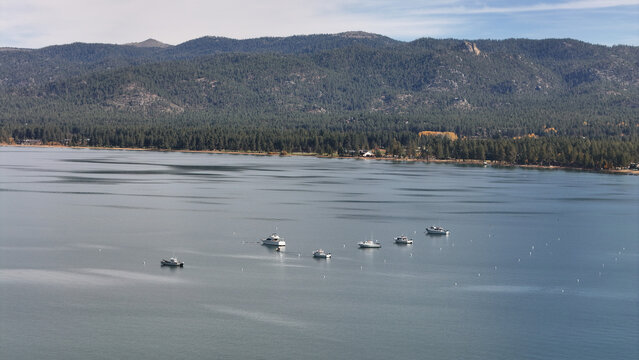 Aerial view of boats gently bobbing on the serene lake under a vast sky, framed by forested mountains, Big Bear Lake, California, United States.