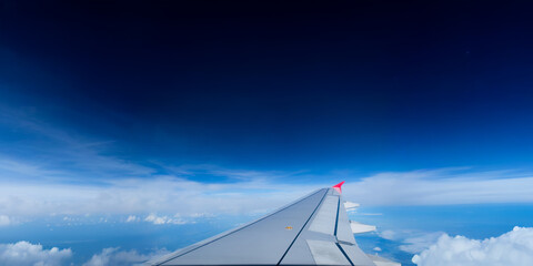 a wing of flying transportation jet plane with a red winglet in deep blue transparent sky background and a cloudy atmosphere below