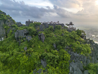 Aerial view of the vibrant green foliage clinging to the rugged, grey cliffs of Hang MÃºa, crowned with a delicate pagoda and arched gateway, Hoa LÆ°, Ninh BÃ¬nh, Vietnam.