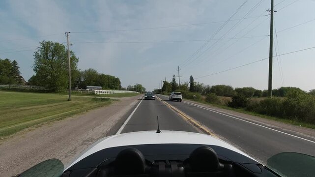Rear POV from a moving convertible, looking down a quiet rural highway. Cars ahead, white fence and fields on both sides. Warm daylight suggests freedom, independence, small town escape.