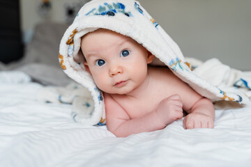 Happy curious baby child after bath with towel on head. Adorable little boy wrapped in blanket. Cutest 3 months old baby smiling in towel on bed close up at home. Child caring routine.