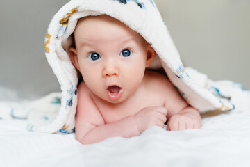 Happy curious baby child after bath with towel on head. Adorable little boy wrapped in blanket. Cutest 3 months old baby smiling in towel on bed close up at home. Child caring routine.