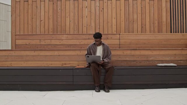 Focused on programming digital nomad sitting on wooden bench in local park, he holding laptop while looking at screen