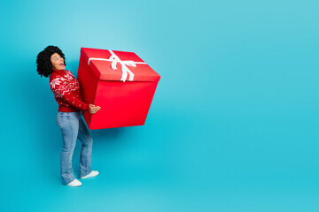 Excited young woman in festive sweater holding a giant red gift box on light blue background