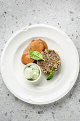 A gourmet dish of meat in a savory sauce, served with a molded grain patty and a side of cucumber salad on a white plate. The food is expertly prepared and presented on a modern speckled tabletop