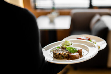 A waiter in a black uniform serves two plates of beautifully plated gourmet food, with a focus on the foreground dish of meat and wild rice. The restaurant setting is softly lit and intimate.